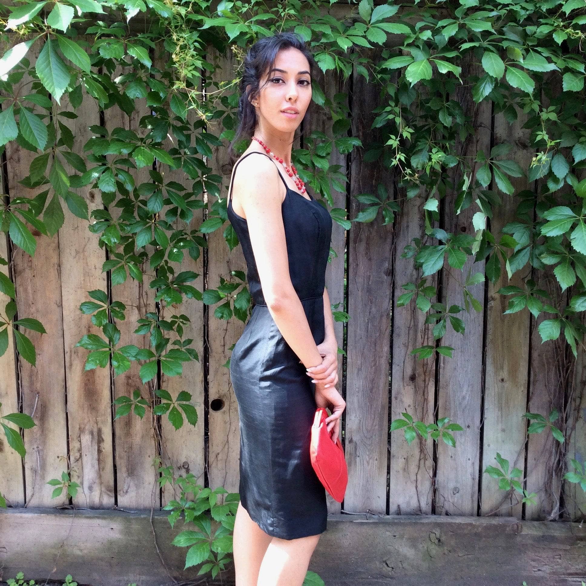 Side View of 1950s Classic Black Silk Cocktail Dress Size Small Model is holding a 1980's Genuine Red Leather Clutch Handbag sold by bohemevintage.com Montreal